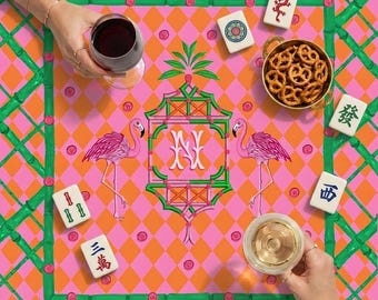 Peut inclure: Une nappe carrée colorée avec un motif de losanges roses et oranges, une bordure verte en forme de bambou et un motif central avec des flamants roses et les lettres "NY". Des tuiles de mahjong et un bol de bretzels sont sur la table, avec des verres de vin rouge et blanc.