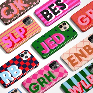 Op de afbeelding: Een verzameling kleurrijke telefoonhoesjes met diverse ontwerpen. Sommige hoesjes tonen initialen in felle kleuren, andere hebben patronen zoals strepen, ruiten en hartjes. De hoesjes zijn ontworpen voor een smartphone.