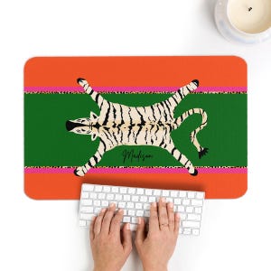 May include: A rectangular desk mat featuring a tiger rug design. The mat has an orange and green background with a pink leopard print stripe. The tiger rug is cream-colored with black stripes, and the name "Madison" is written in cursive.
