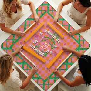 May include: A square mahjong mat featuring a pink and orange checkered pattern with green bamboo-style borders and illustrations of pink flamingos. The mat displays a central monogram and scattered white mahjong tiles with black characters.