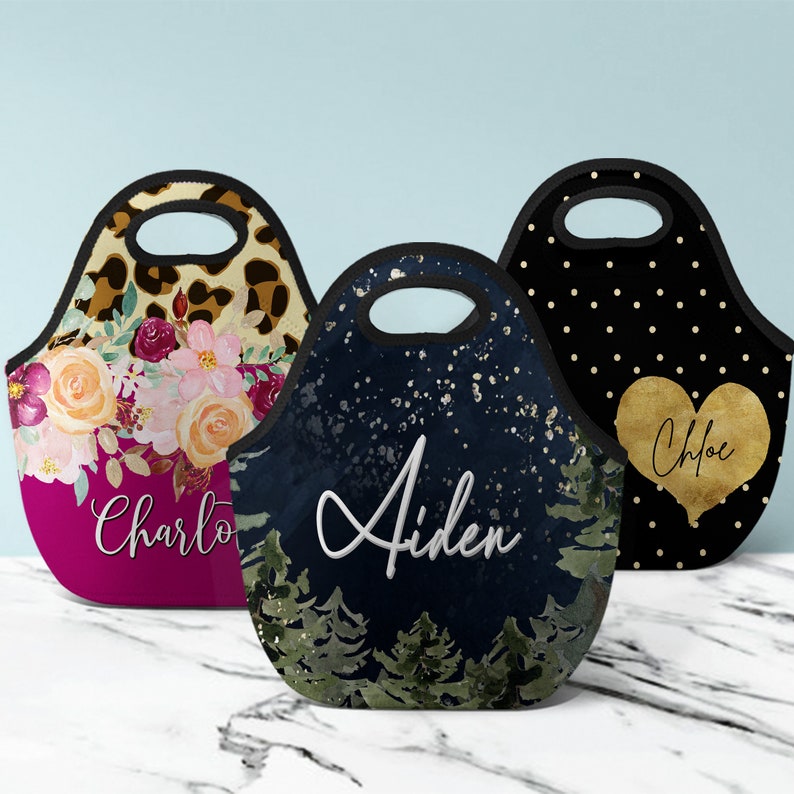 Puede incluir: Tres bolsas de almuerzo personalizadas con asas negras. Una bolsa presenta un estampado de leopardo y un dise&ntilde;o floral con el nombre "Charlotte". Otra tiene una escena de noche estrellada con el nombre "Aiden". La tercera bolsa tiene un fondo negro con lunares dorados y un coraz&oacute;n dorado con el nombre "Chloe".