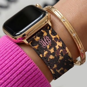 May include: Gold Apple Watch with a tortoiseshell patterned strap and a pink monogram. A gold bracelet with small diamonds is also visible. The watch is on a wrist with a pink sweater.
