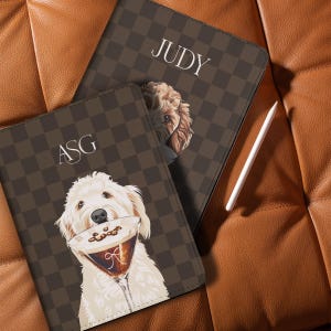 May include: Two brown and tan personalized tablet cases with dog illustrations. One case has the initials "ASG" and a dog holding a cocktail. The other case has the name "JUDY" and a dog's face. A white stylus is next to the cases.
