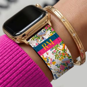 May include: Gold Apple Watch with a floral patterned strap. The strap has a white background with pink, yellow, and blue floral designs, a teal stripe, a pink stripe, and the initials "RM" in gold.