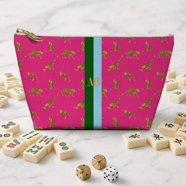May include: A pink zippered pouch with a gold leopard print pattern. The pouch has a green and light blue stripe with the gold letters "AB". Mahjong tiles and dice are in the foreground.