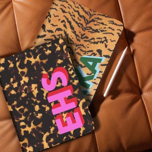 May include: Two patterned tablet cases. One case has a tortoiseshell design with the letters "EHS" in pink and red. The other case has a tiger stripe pattern with the letters "AKA" in green and blue. A white stylus is also visible.