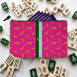 May include: A pink zippered pouch with a gold leopard print and a green and light blue stripe. The pouch has gold initials "AB" and is surrounded by Mahjong tiles.