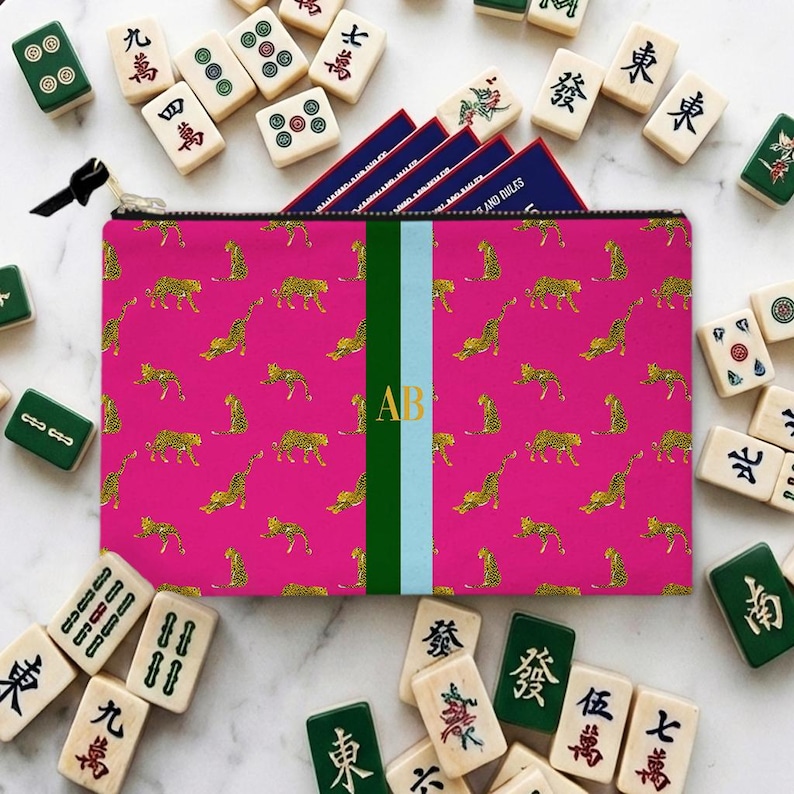 May include: A pink zippered pouch with a gold leopard print and a green and light blue stripe. The pouch has gold initials "AB" and is surrounded by Mahjong tiles.
