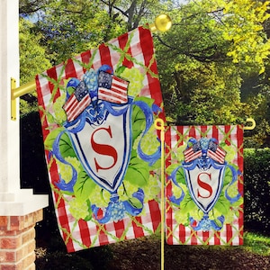 May include: A pair of red, white, and blue garden flags with a white shield featuring the letter "S" in red. The flags are decorated with blue ribbons, green leaves, and white flowers. The flags are on a red and white checkered background.