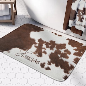 May include: A brown and white cowhide patterned bath mat with the name "Harrison" written in cursive on it.
