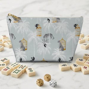 Peut inclure: Trousse de maquillage bleu clair avec fermeture &eacute;clair dor&eacute;e, orn&eacute;e d'un motif de l&eacute;opards, de singes et de cages &agrave; oiseaux. Des tuiles de mahjong et des d&eacute;s entourent le sac. Les initiales "SME" sont imprim&eacute;es.