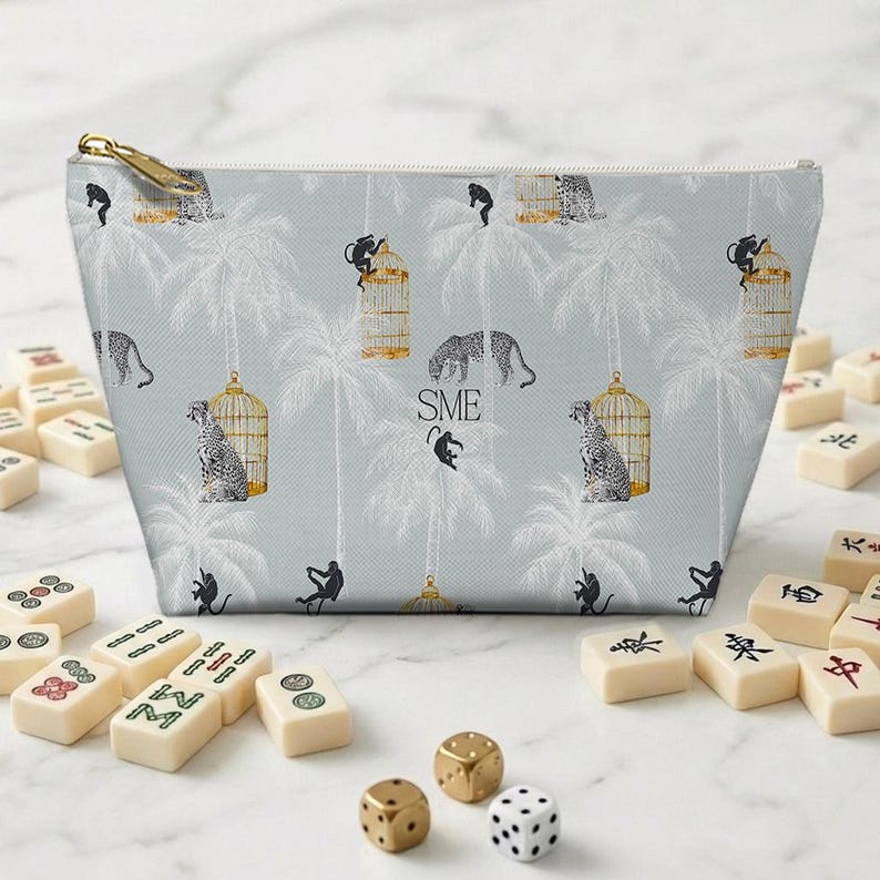 Peut inclure: Trousse de maquillage bleu clair avec fermeture &eacute;clair dor&eacute;e, orn&eacute;e d'un motif de l&eacute;opards, de singes et de cages &agrave; oiseaux. Des tuiles de mahjong et des d&eacute;s entourent le sac. Les initiales "SME" sont imprim&eacute;es.