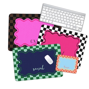 May include: A collection of personalized mouse pads in various designs. The pads feature a pink, blue, and orange scalloped border with names and initials. One pad has a brown and black checkered pattern, and another has a black and white checkered pattern. A silver keyboard is also present.