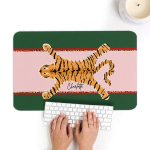 May include: A rectangular desk mat featuring a stylized orange and black tiger illustration on a pink background. The mat has a green border with a leopard print stripe. The name "Charlotte" is written in cursive. A white keyboard is in the foreground.