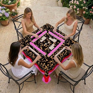 May include: A square table covered with a leopard print tablecloth. Four people are seated around the table, playing a game of Mahjong. The Mahjong tiles are white with black characters, and the racks are bright pink.