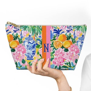 May include: A zippered cosmetic bag with a floral pattern featuring oranges, pink hydrangeas, and blue and white vases. The bag has an orange and pink stripe with the letter "N".