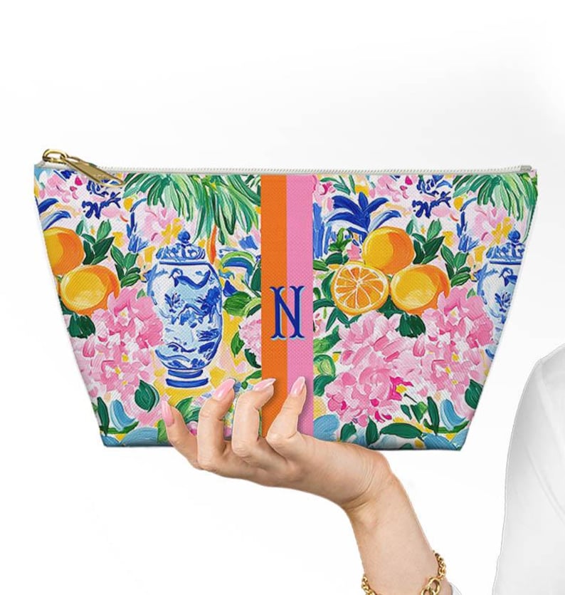 May include: A floral print cosmetic bag with a gold zipper, featuring oranges, pink hydrangeas, and blue and white vases. An orange and pink stripe with the letter "N" is centered on the bag.