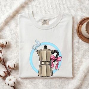 May include: A white t-shirt featuring a graphic of a silver moka pot with a pink bow tied to the handle and a wisp of blue steam rising from the top, all centred within a light blue circle. The shirt has a Comfort Colors brand neck label.