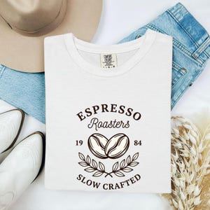 May include: A white short-sleeved t-shirt featuring a dark brown graphic print that reads Espresso Roasters 1984 Slow Crafted, centred above and below two coffee beans and a decorative leaf branch design, with a Comfort Colors brand tag.