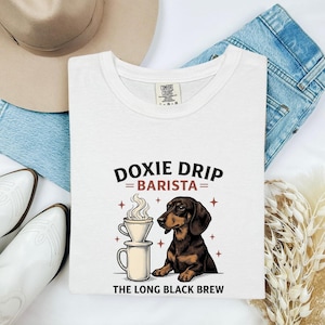 May include: A white t-shirt featuring a graphic of a brown dachshund sitting next to a coffee dripper and mug. The text reads DOXIE DRIP BARISTA and THE LONG BLACK BREW. The shirt has a Comfort Colors label at the inner neckline.