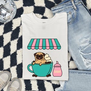 May include: A white t-shirt featuring a graphic of a tan pug sitting inside a large teal coffee cup topped with whipped cream. Above the cup is a pink and teal striped awning, and next to it is a pink sugar shaker with the word SUGAR printed on it.