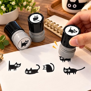 May include: Three cylindrical self-inking stamps with black caps and grey bases. Each stamp features a white circular top label displaying a unique black cat illustration. One stamp is shown in use, creating black cat imprints on white paper.