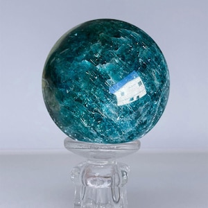 May include: A polished, spherical blue-green mineral stone with visible internal crystalline fractures and light reflections, resting on a clear, transparent glass stand with a flared top and a sturdy, multi-faceted base.