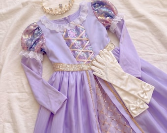 May include: A lavender dress with long sleeves, puff shoulders featuring iridescent sequins, a white lace collar, and a bodice with crisscross ribbon detailing. The set includes a gold-toned metal tiara with clear crystals and white satin gloves.
