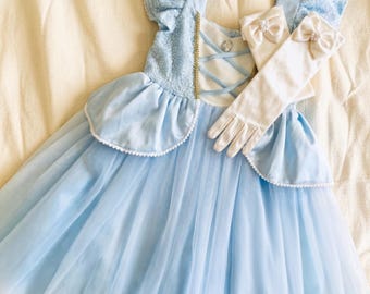 May include: A light blue dress with a textured bodice, ruffled sleeves, and a layered tulle skirt. The bodice features a white center panel with blue crisscross lacing and gold trim. A pair of white satin gloves with small bows and pearls is included.