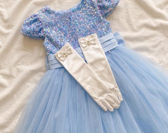 May include: A light blue dress featuring a bodice covered in iridescent sequins, short puff sleeves, and a layered tulle skirt. A wide satin waistband sits at the waist. A pair of white satin gloves with small bows and faux pearls are placed on top.