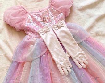 May include: A pink dress with puff sleeves, a sequined bodice featuring a central snowflake emblem, and a multi-colored tulle skirt with silver star and moon patterns. A pair of matching pale pink satin gloves with small bows and pearls.