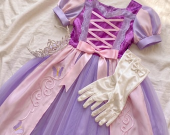 May include: A purple and pink dress with puff sleeves, a lace-trimmed bodice featuring a crisscross ribbon design, and a tulle skirt adorned with embroidered butterfly motifs. The set includes a silver-toned tiara and white satin gloves.