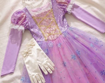 May include: A purple long-sleeve dress featuring a sequined bodice with a gold crisscross pattern, puffed sleeves with white lace trim, and a layered tulle skirt adorned with blue floral appliqués, accompanied by a silver tiara and white gloves.