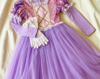 May include: A long-sleeved dress featuring a pink sequined bodice with a gold crisscross pattern, purple puff sleeves with white lace trim, and a full-length lavender tulle skirt. A pair of white gloves with small bows is placed on the dress.