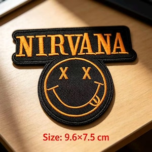 May include: A black embroidered patch featuring the word NIRVANA in orange capital letters above a circular smiley face icon with X-shaped eyes and a tongue sticking out. The patch measures 9.6 by 7.5 cm as indicated by the text below.