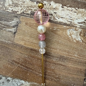 May include: A decorative metal pen featuring a series of stacked beads in varying sizes and textures. The beads include a large faceted pink bead, a smooth white pearl-like bead, a pink glitter bead, a silver glitter bead, and a clear bead.