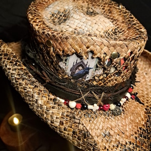May include: A brown woven straw hat featuring a dark leather band adorned with a string of beads, including small white crosses, red roses, and black spheres. A rectangular patch displays a black raven perched before a gothic arched window.
