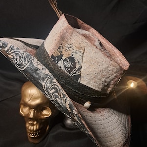 May include: A beige straw hat with a black band, a feather accent, and a black and white patterned brim. The hat features a rectangular patch with a dark graphic design on the side and is positioned next to a metallic gold skull figurine.