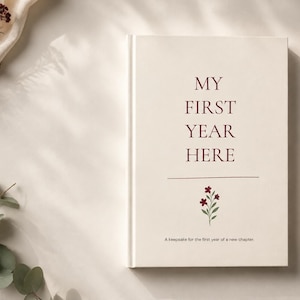 Può includere: Un libro con copertina rigida color crema con il testo "MY FIRST YEAR HERE" stampato in rosso scuro, caratterizzato da una piccola illustrazione floreale e dal sottotitolo "A keepsake for the first year of a new chapter" sotto una linea orizzontale, accompagnato da una penna dorata.