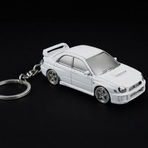 Puede incluir: Un llavero de coche sedán en miniatura de color blanco con alerón trasero, ruedas plateadas y ventanas de tono plateado. El modelo del vehículo incluye una toma de aire en el capó, espejos laterales y una anilla metálica.