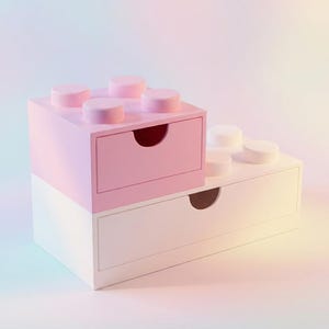 May include: A two-tiered storage unit shaped like interlocking building blocks. The top section is a pink square drawer with four circular studs on top. The bottom section is a wider white rectangular drawer with two circular studs on top.