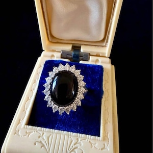 May include: A ring featuring a large, oval-shaped black stone set in a silver-toned metal band, surrounded by a halo of small, clear, sparkling stones. The ring is displayed inside an open, cream-colored jewelry box with a blue velvet lining.
