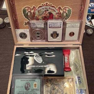 Puede incluir: Una caja de puros de madera que contiene una colección de artículos, incluyendo tres monedas encapsuladas, un estuche negro de la United States Mint, una barra de KitKat, una tarjeta dorada American Success, un billete Goldback y un billete polaco.