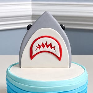 May include: A shark cake topper featuring a grey triangular body, two small black eyes, and a white mouth area outlined in red with jagged red teeth. The topper sits atop a round cake with layered light blue and white frosting ruffles.