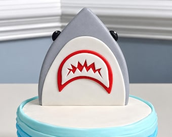 May include: A shark cake topper featuring a grey triangular body, two small black eyes, and a white mouth area outlined in red with jagged red teeth. The topper sits atop a round cake with layered light blue and white frosting ruffles.