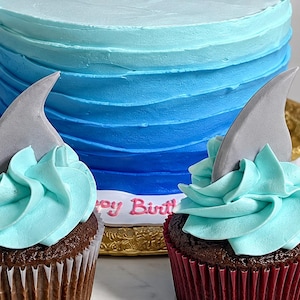 May include: A round cake with blue ombre frosting and a white base border featuring the text Happy Birthday in pink, accompanied by two chocolate cupcakes topped with light blue swirled frosting and gray shark fin decorations.