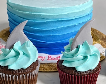 May include: A round cake with blue ombre frosting and a white base border featuring the text Happy Birthday in pink, accompanied by two chocolate cupcakes topped with light blue swirled frosting and gray shark fin decorations.