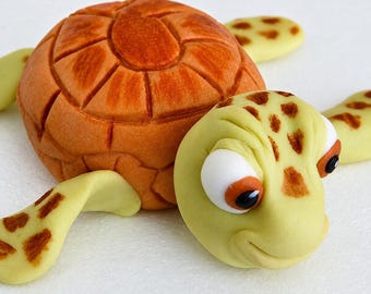 May include: A sculpted turtle figurine made of smooth material, featuring a rounded, textured brown shell with defined segments and a light yellow head and flippers adorned with brown spots. The turtle has large white eyes with dark pupils.