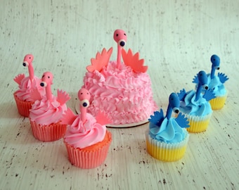 Op de afbeelding: Een kleine roze taart met roze glazuur en een flamingo-figuurtje, omringd door drie roze cupcakes met flamingo-toppers en drie blauwe cupcakes met blauwe flamingo-toppers, allemaal voorzien van gegolfd glazuur en papieren vormpjes.