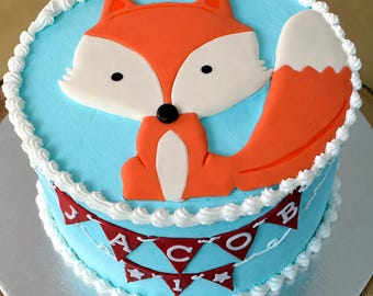 Op de afbeelding: Een ronde taart met lichtblauw glazuur en een witte rand. De bovenkant heeft een platte oranje en witte vosdecoratie. De zijkant van de taart toont een rode vlaggenlijn met witte letters die de naam JACOB spellen en kleine sterretjes.